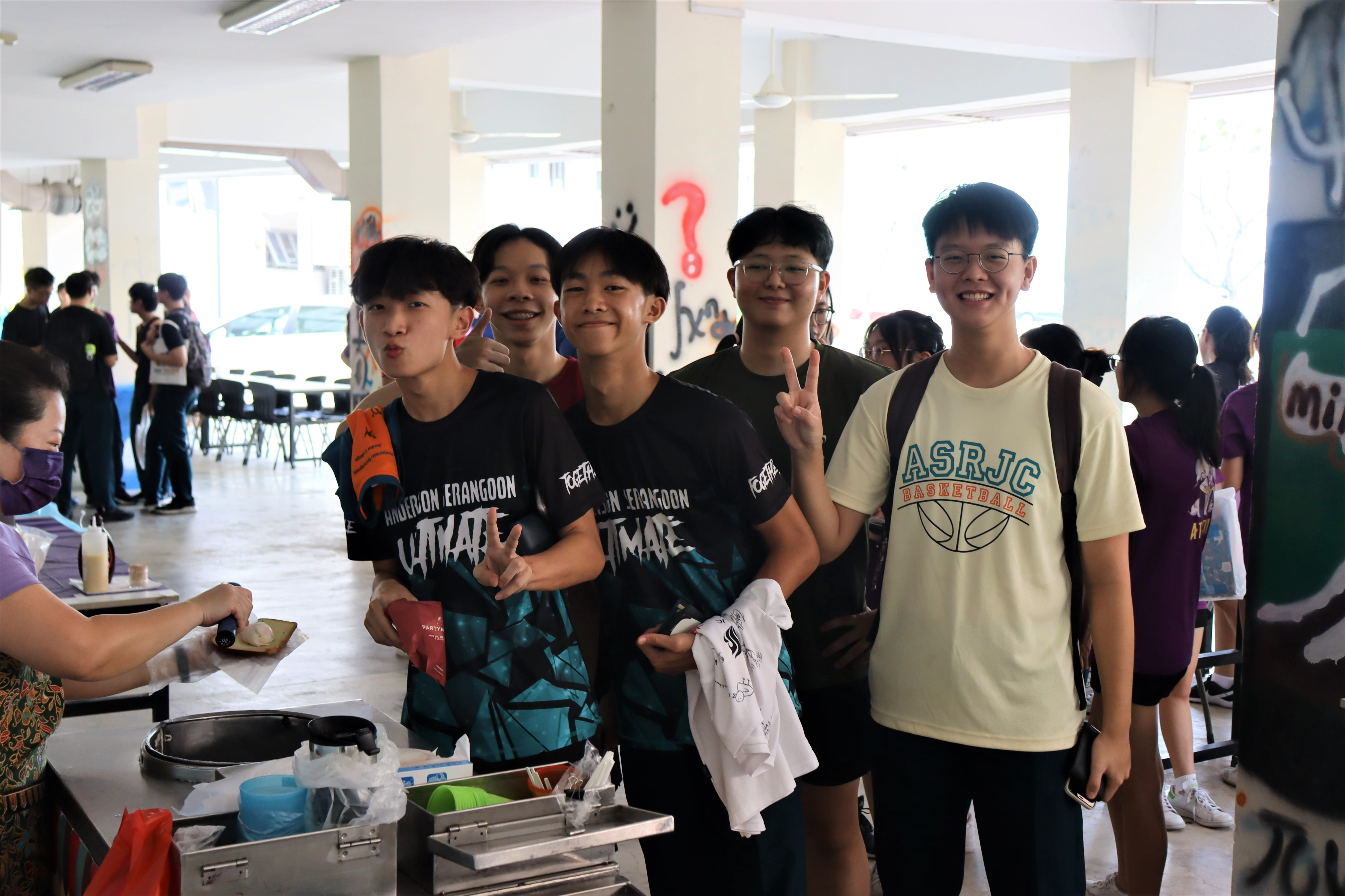 Five smiling young men pose together at an indoor food stall, some making peace signs, wearing sports jerseys.